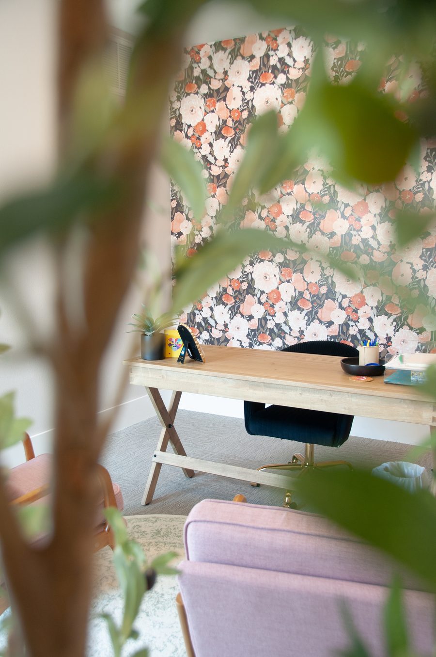 A calm, organized home office with a wooden desk, navy chair, and floral wallpaper